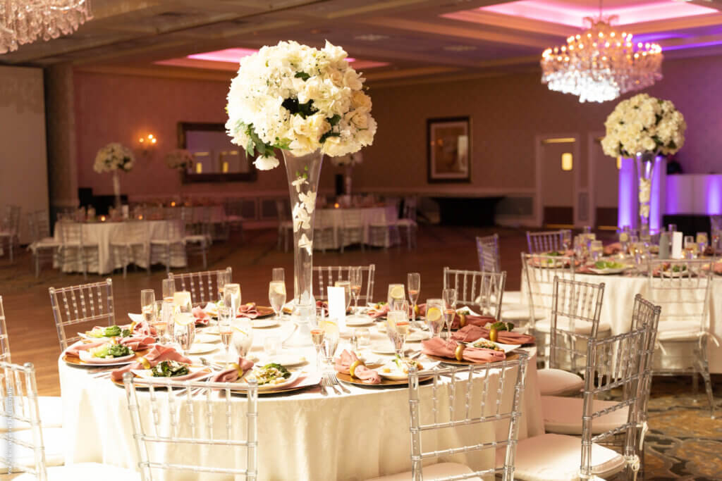 the balrrom set with white tablecloths, pink napkins, floral centerpieces and pink lighting.