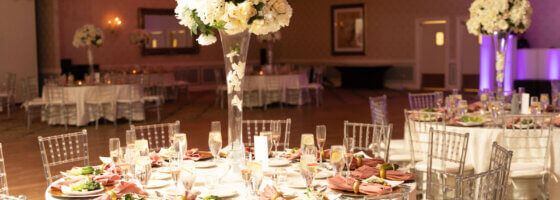 the balrrom set with white tablecloths, pink napkins, floral centerpieces and pink lighting.